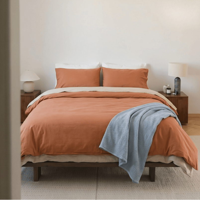 A neatly made bed in a minimalist bedroom, styled with an orange duvet cover, matching orange pillows, a light beige sheet, and a light blue throw blanket. The room features a white wall, a hanging pendant light, wooden bedside tables, and a light-colored rug.
