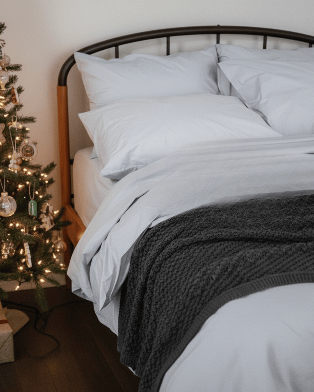 Bed with white bedding and a dark gray throw, next to a decorated Christmas tree.